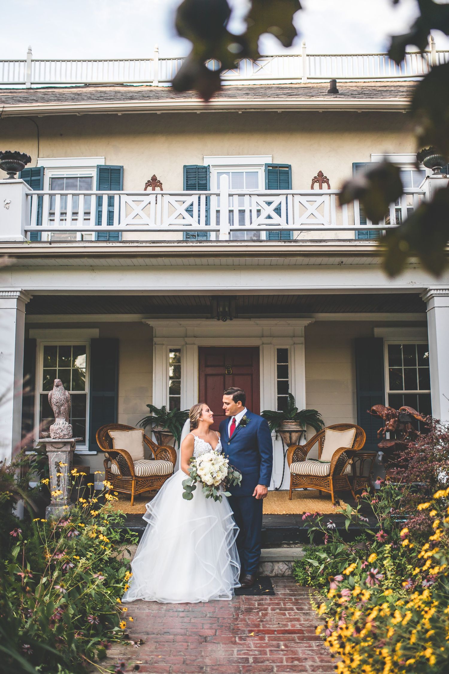 A couple poses on a historic home's front porch surrounded by yellow flowers and classic architecture.