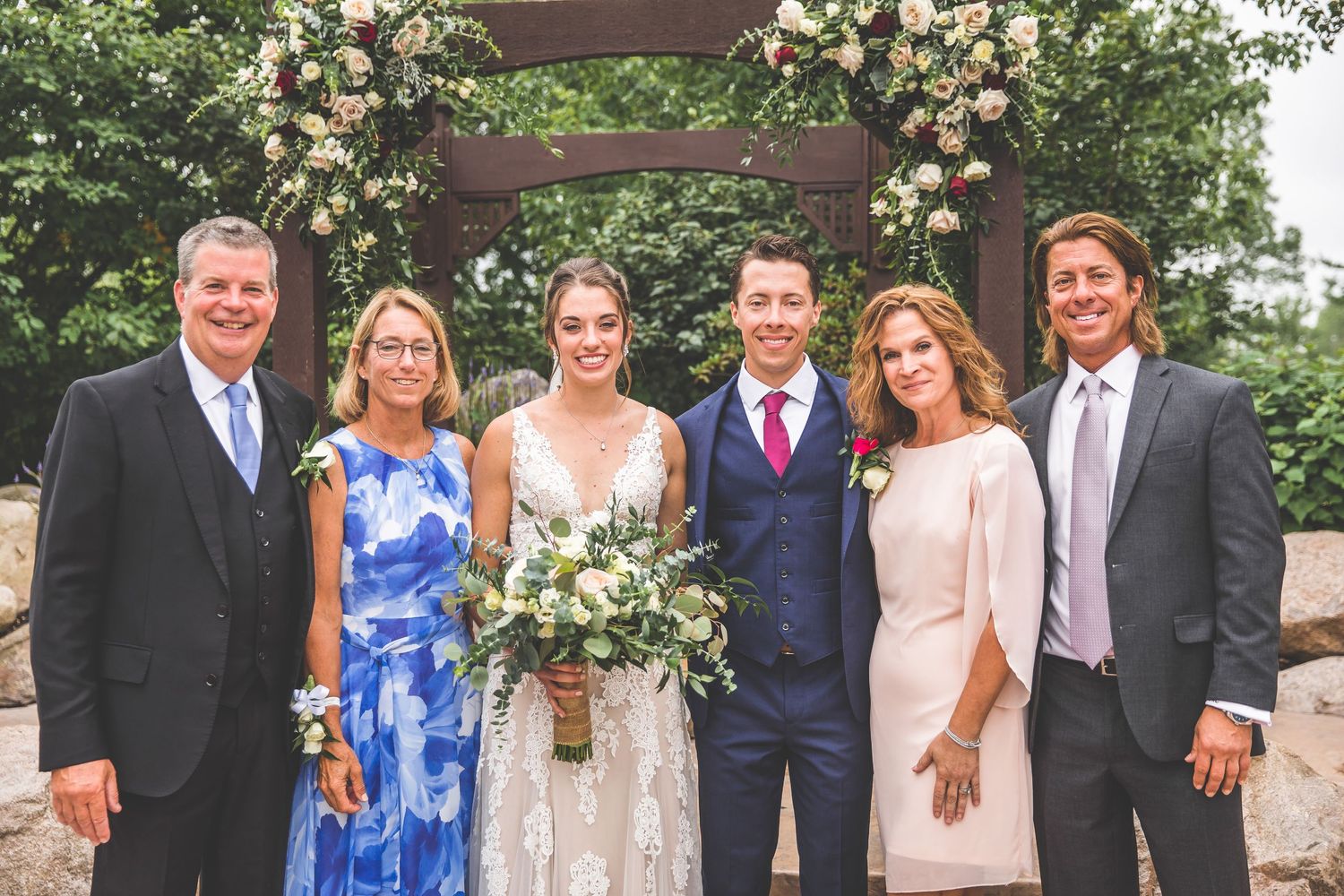 Wedding party members pose in blue dresses and formal suits under floral arch.