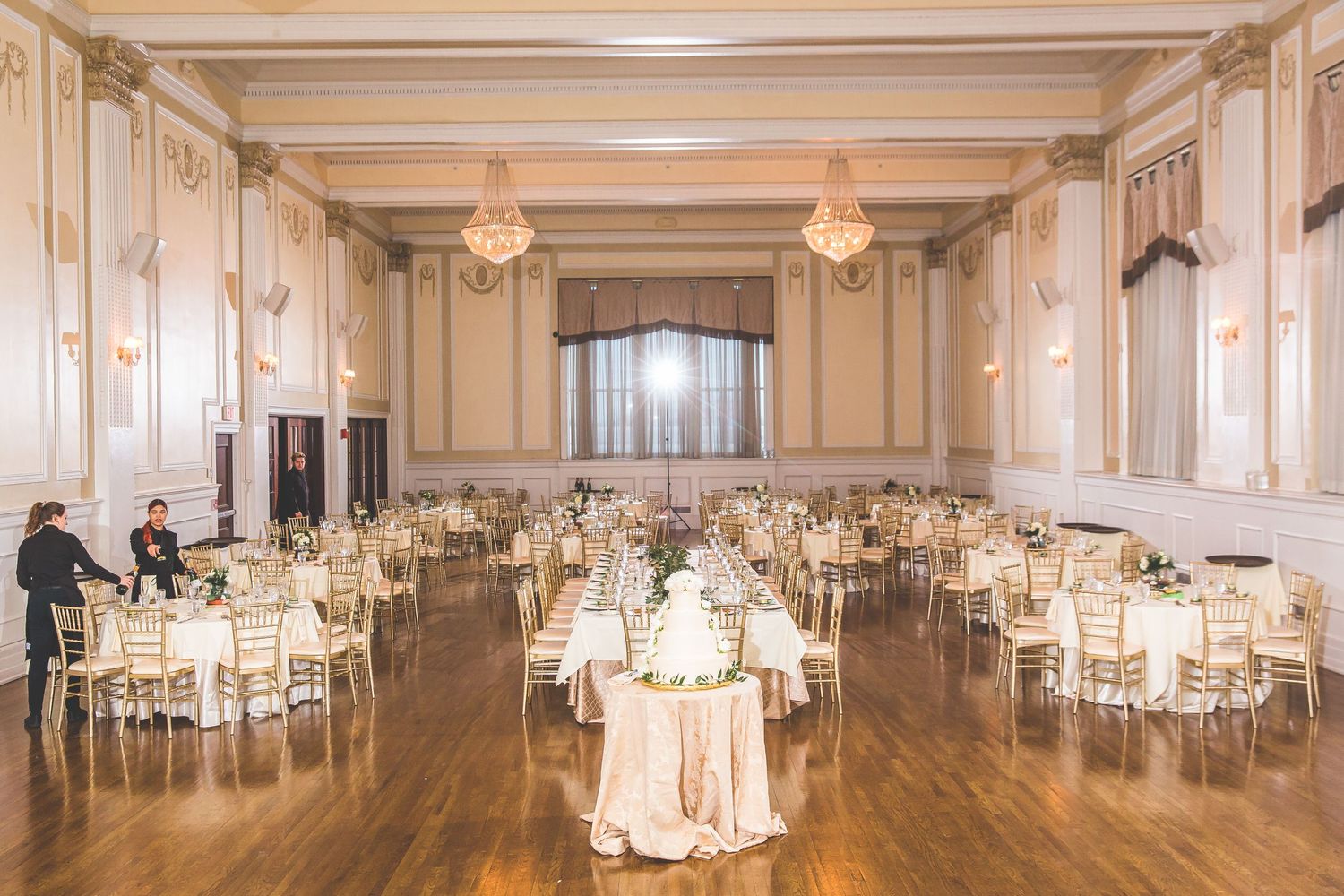Elegant ballroom set for a wedding reception with round tables and warm lighting under cream-colored ceiling.