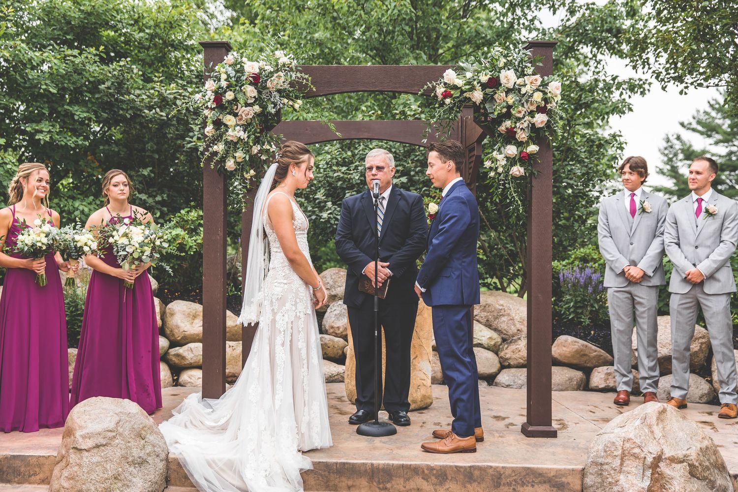 Wedding party members lined up during outdoor ceremony with floral decorations and stone columns.