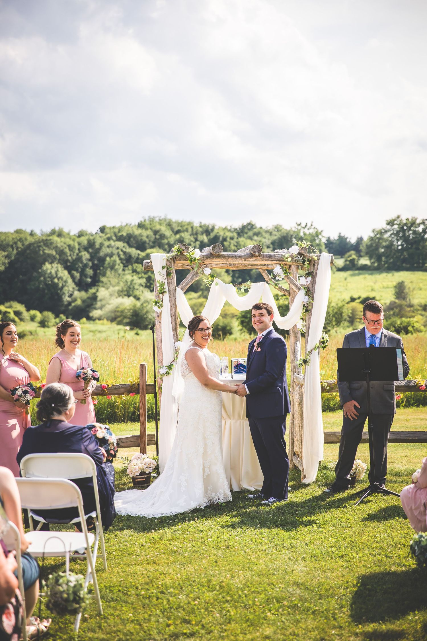 An outdoor wedding ceremony with attendants in pink dresses gathered around a wooden arch decorated with white fabric.