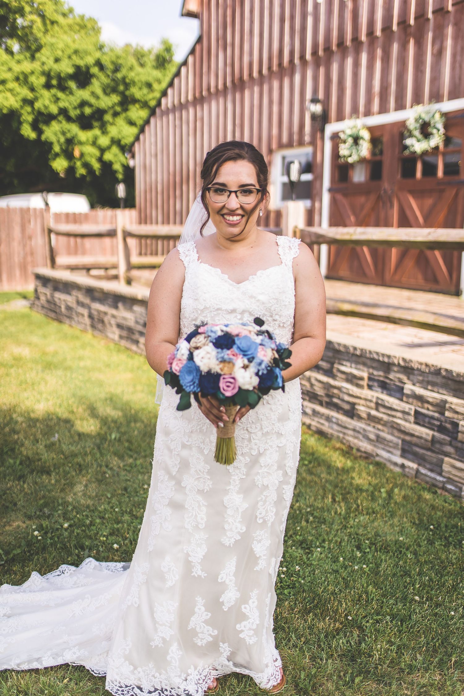 White lace wedding dress with floral bouquet photographed against rustic barn backdrop.