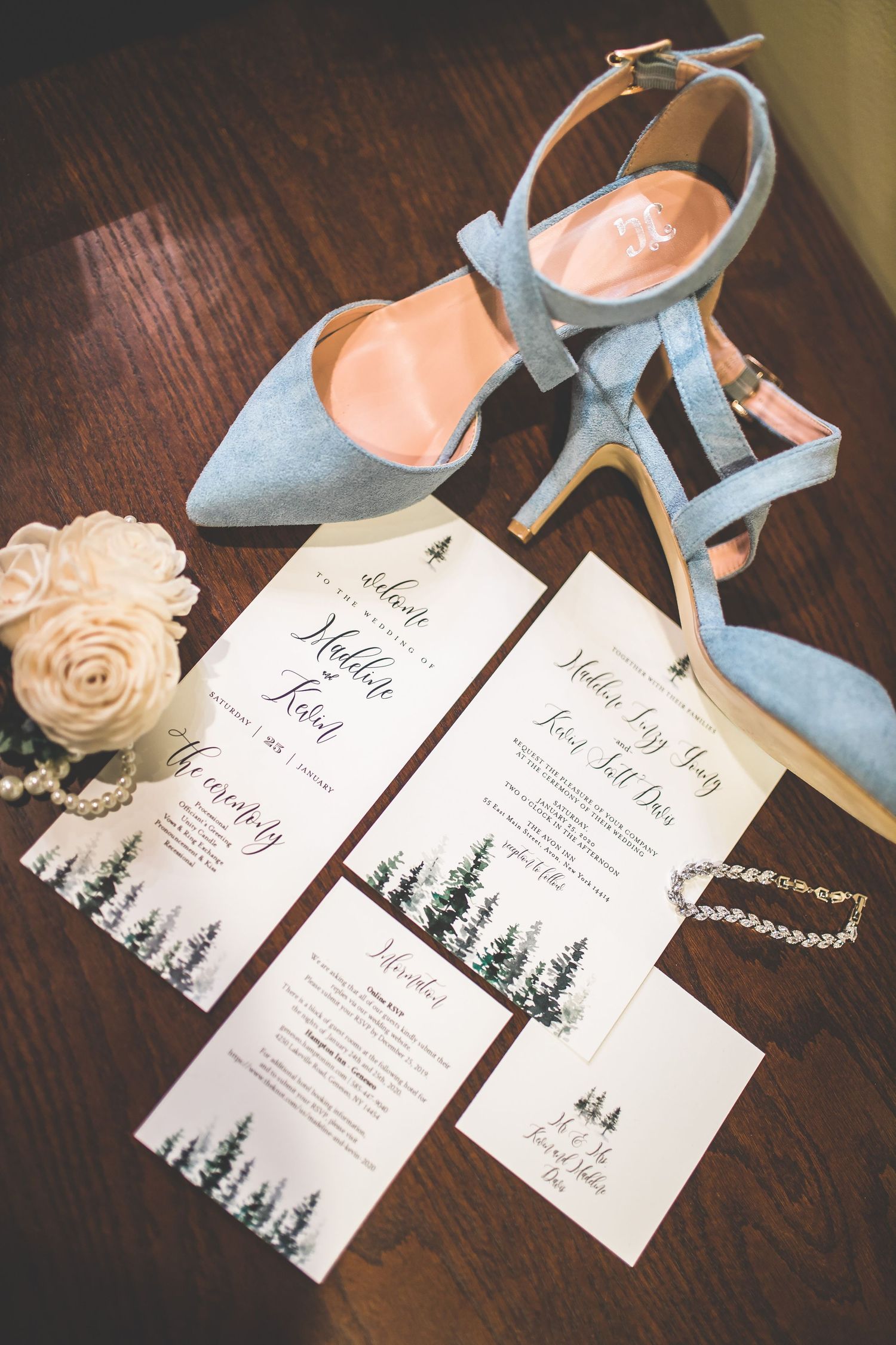 Wedding invitations with forest designs and blue shoes displayed on a dark wooden surface.