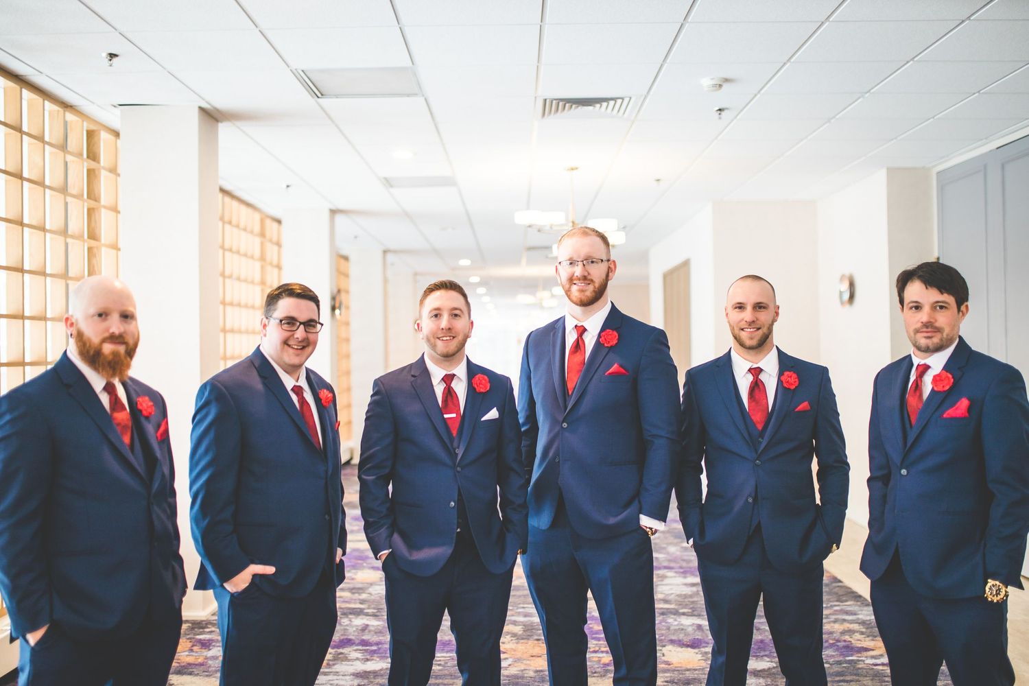 Groomsmen in navy suits with red ties pose together in modern indoor setting.