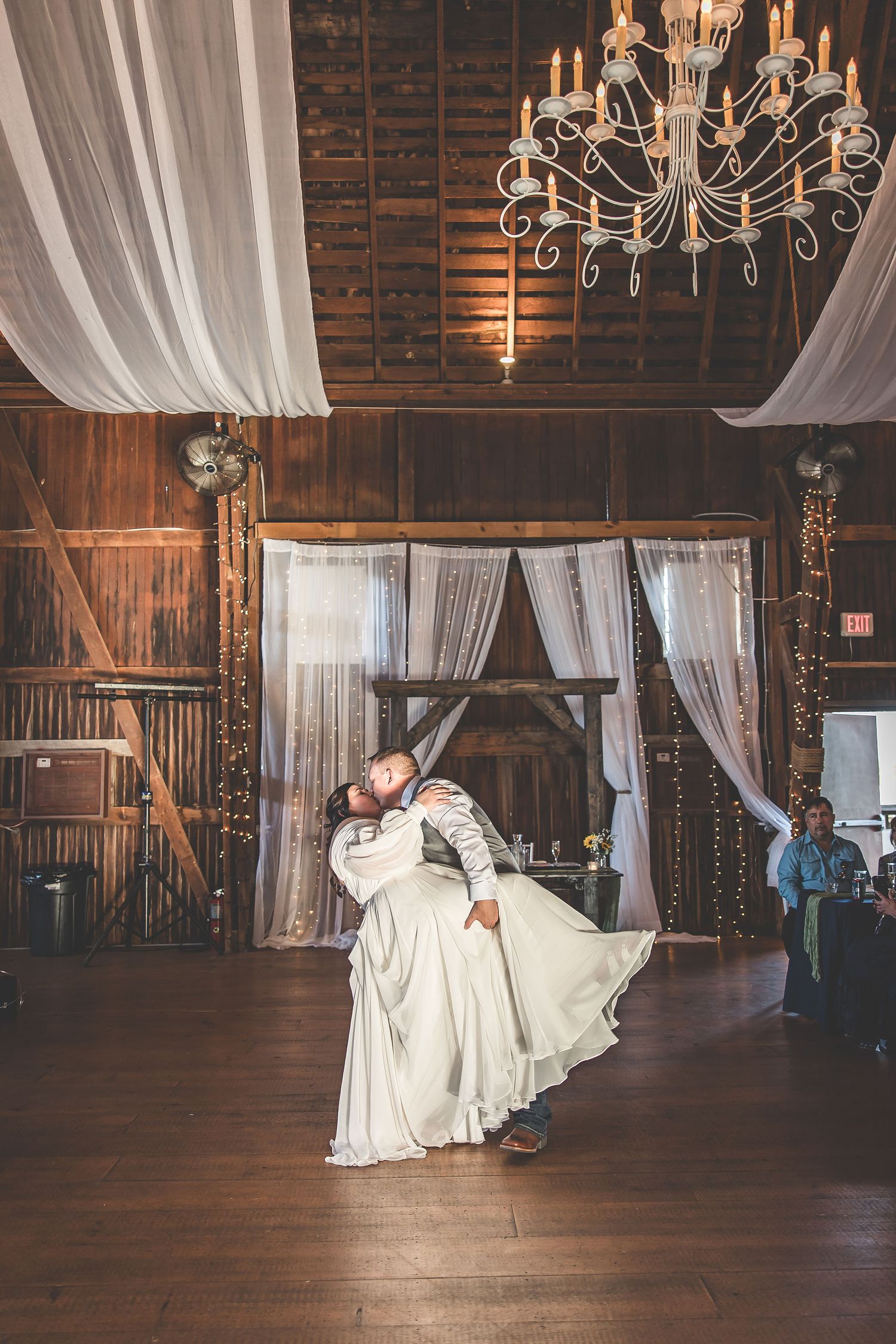 Wedding dance celebration in a rustic barn venue with elegant chandeliers and white draping.