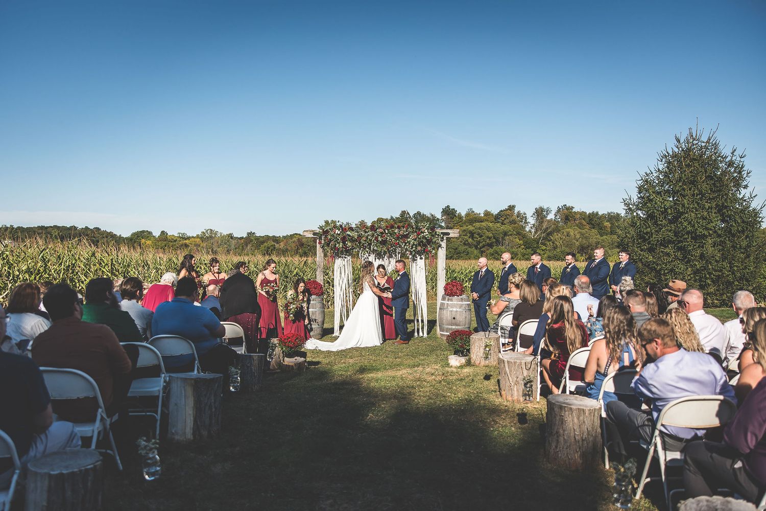 Wedding guests seated in white chairs on a grassy field during an outdoor ceremony.