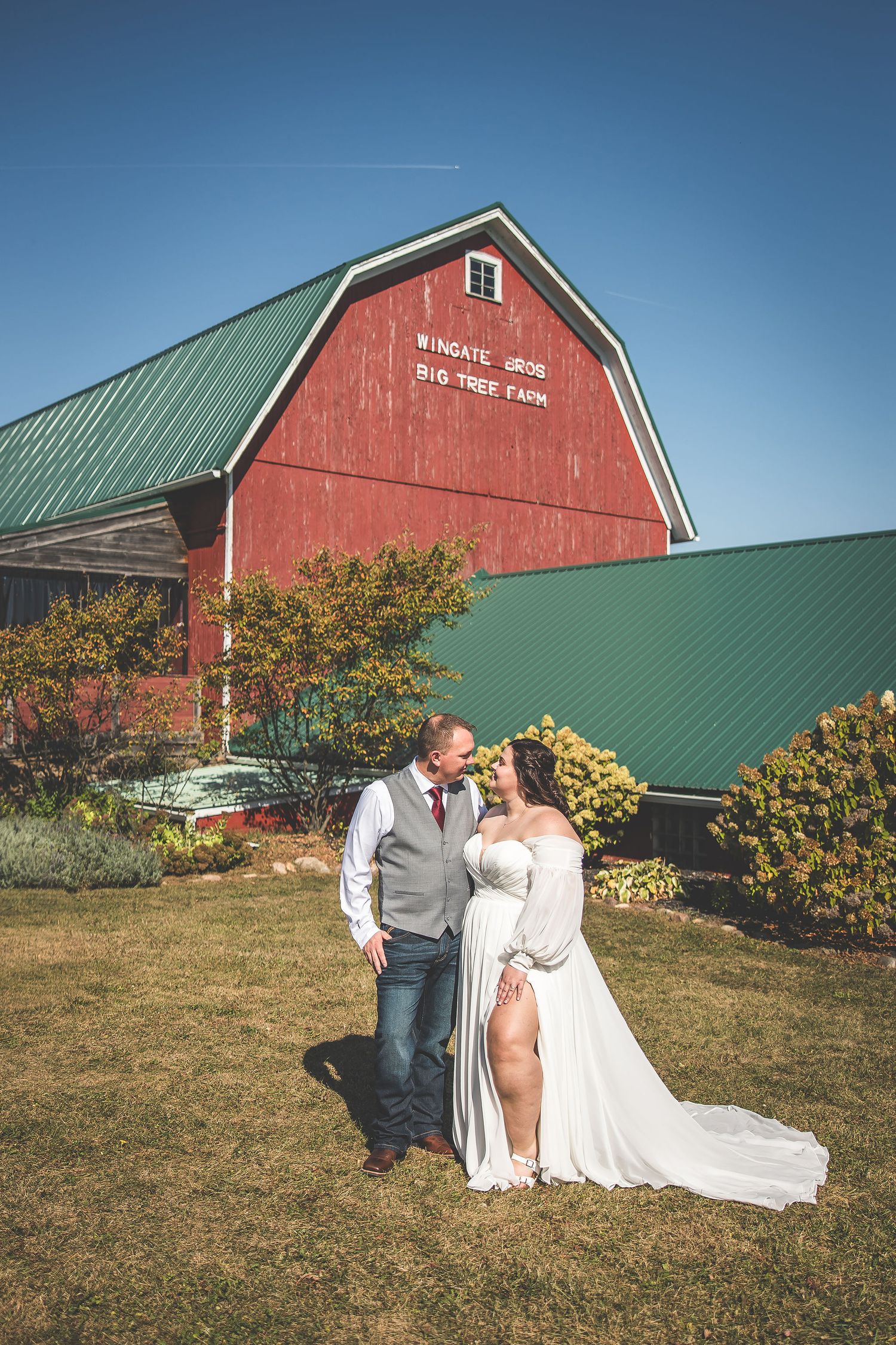 Wedding couple poses in front of red barn with green roof on sunny autumn day.