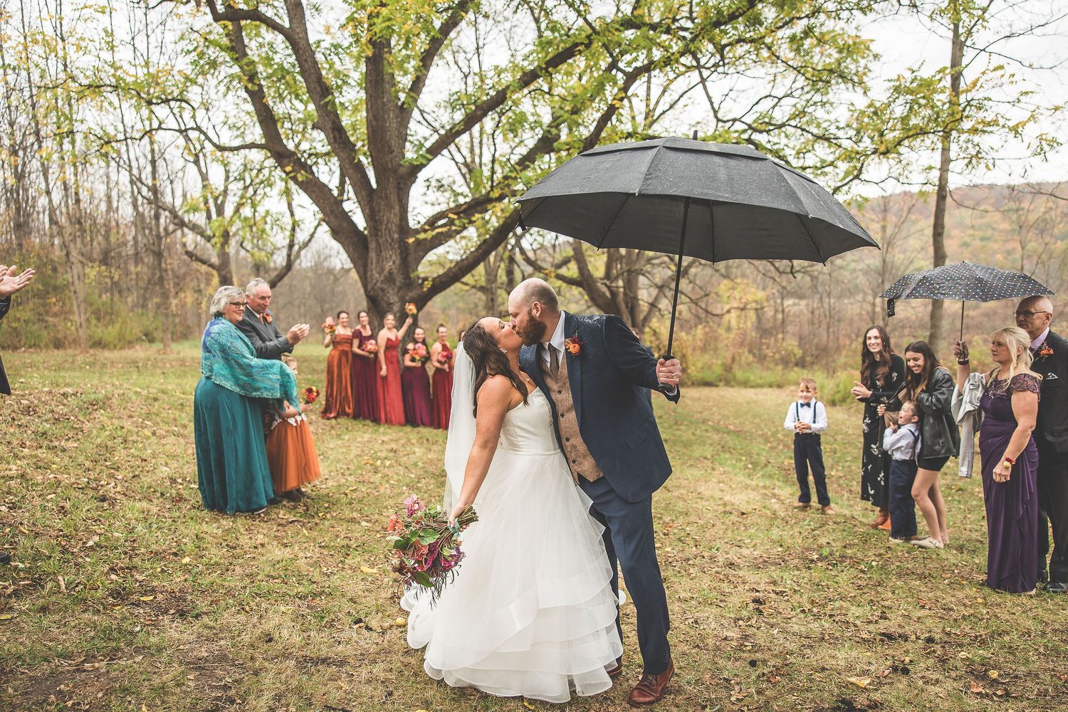 Wedding celebration in a rustic outdoor setting with umbrellas and fall foliage.