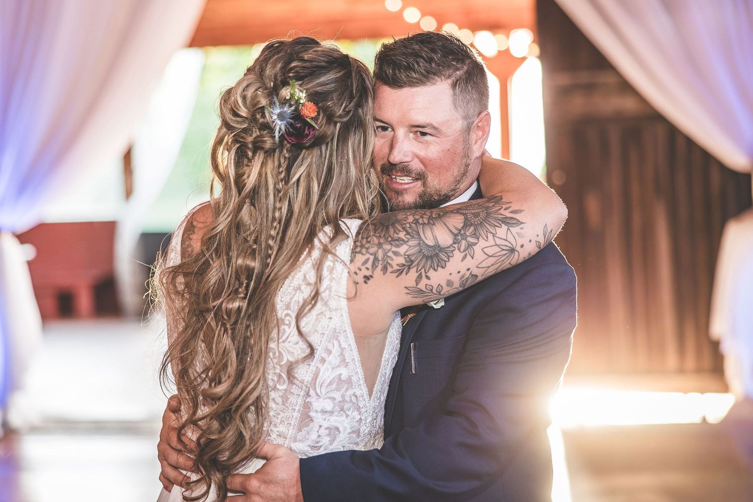 Romantic dance moment captured at rustic barn wedding with soft purple lighting and white drapery.