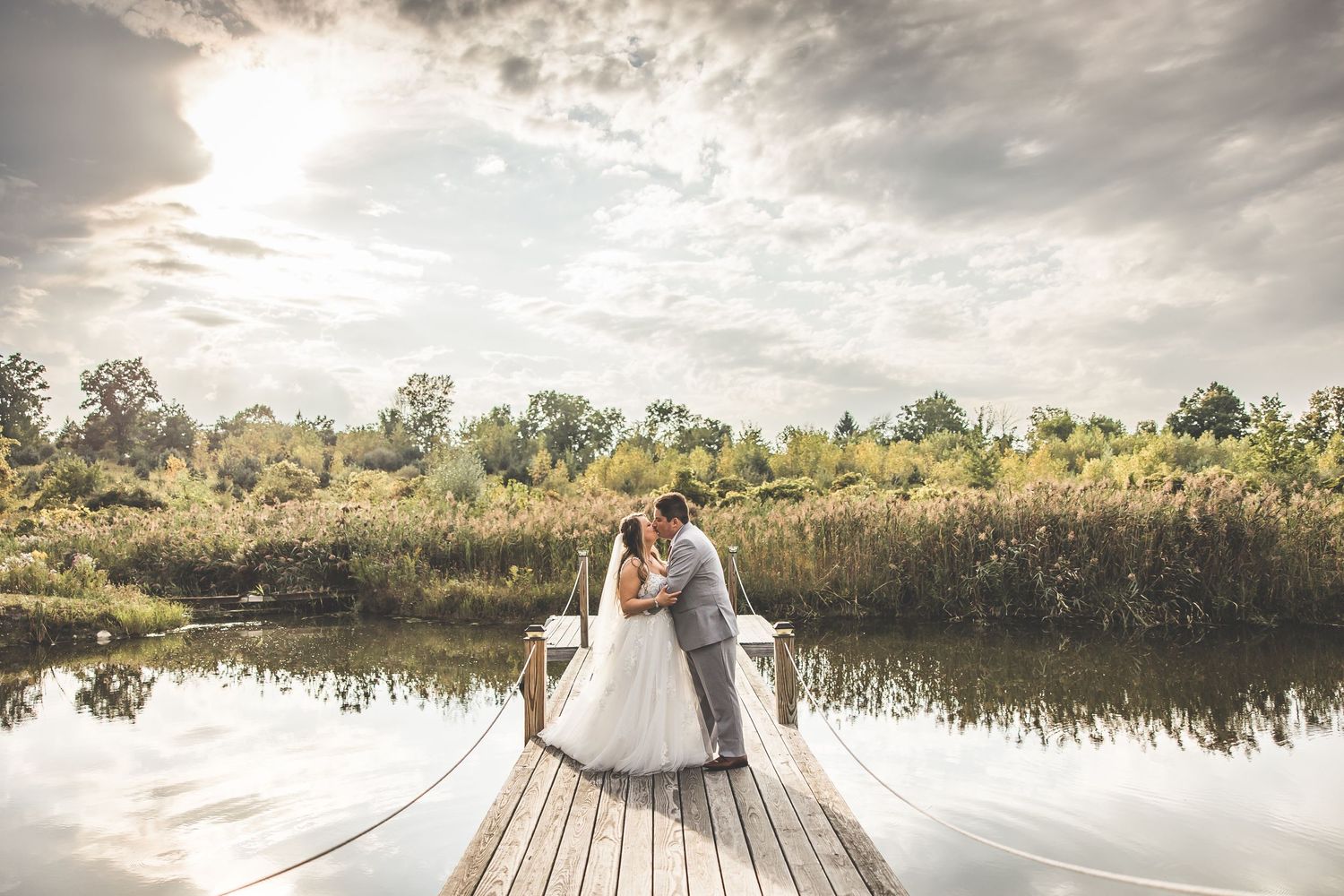 A scenic wedding photo shoot takes place on a wooden dock with dramatic cloudy skies overhead.