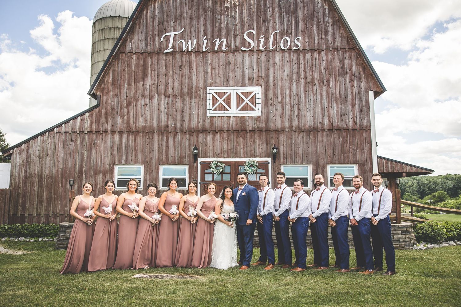Wedding party poses in front of a rustic brown barn on a sunny day.
