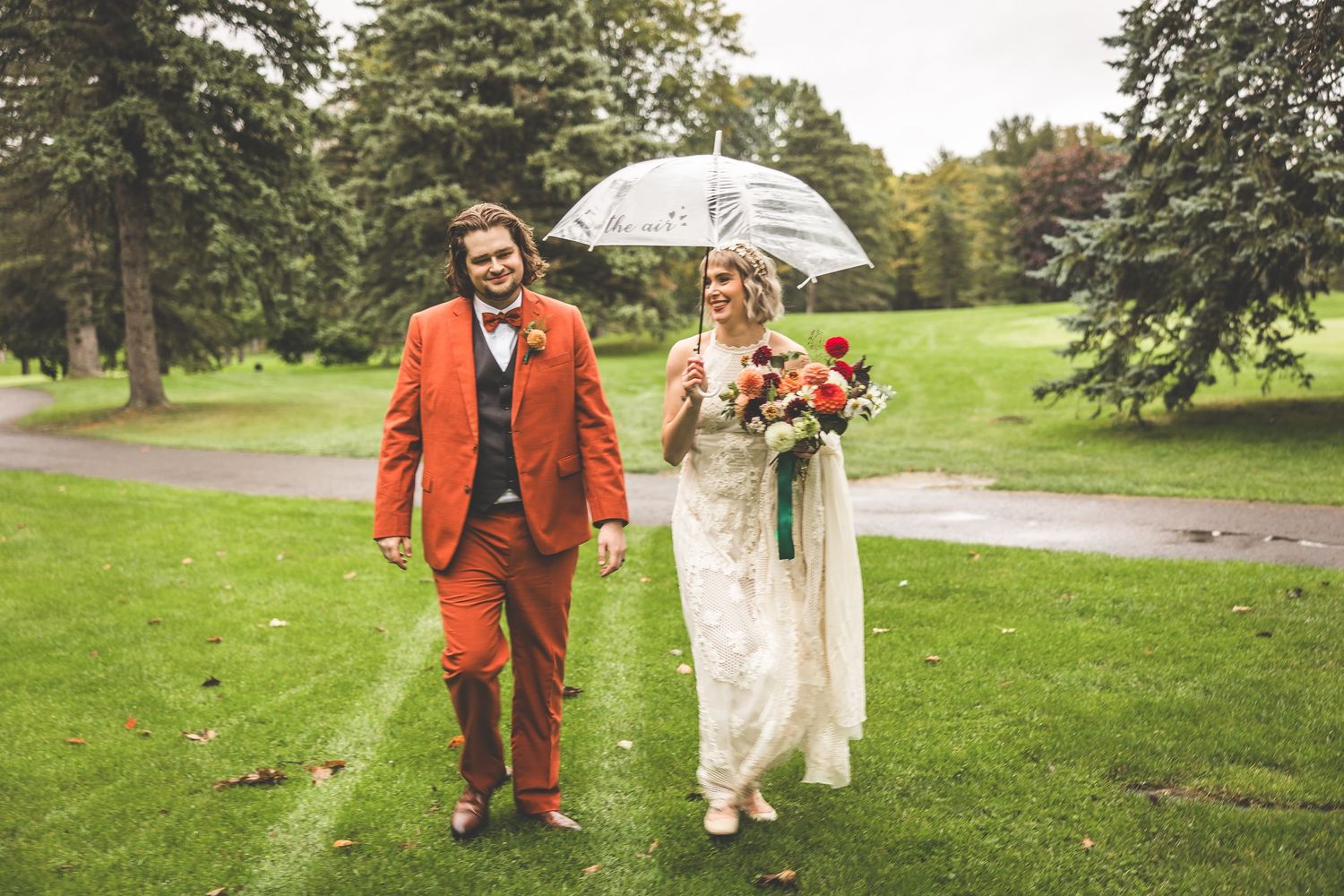 Wedding couple walks together holding a clear umbrella in a lush garden setting.