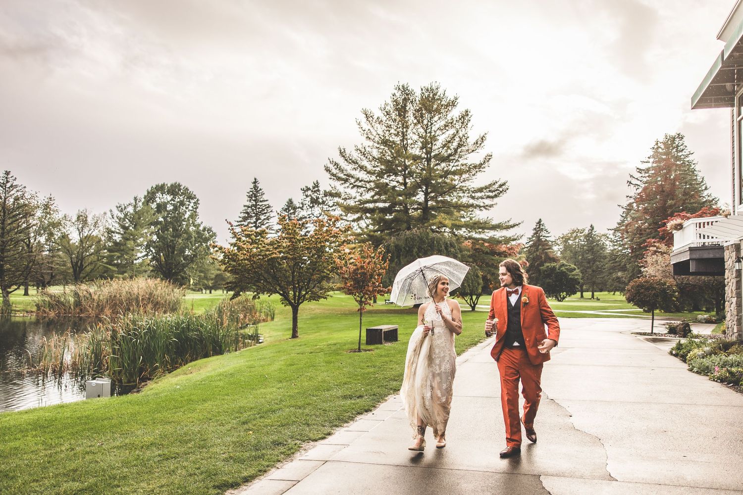 Couple walking along wet pathway with umbrella against cloudy sky and pine trees.