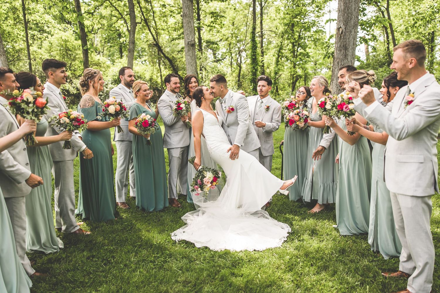 Wedding party forms a circle around the couple on a sunny lawn.
