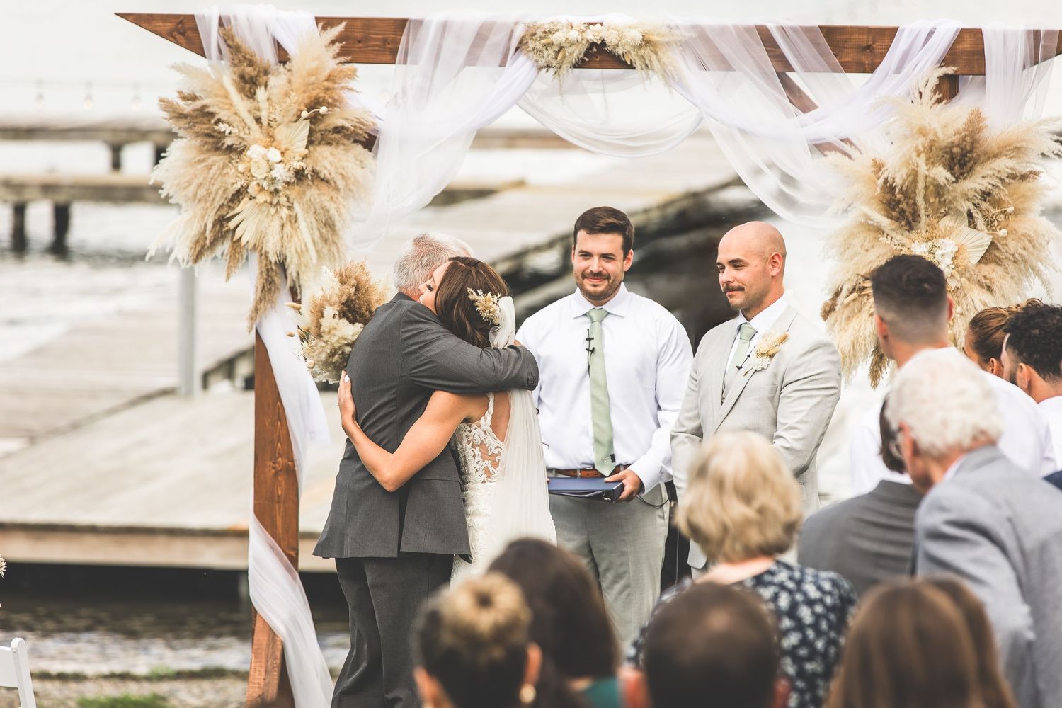 Wedding party members celebrate with pampas grass bouquets against a waterfront backdrop.