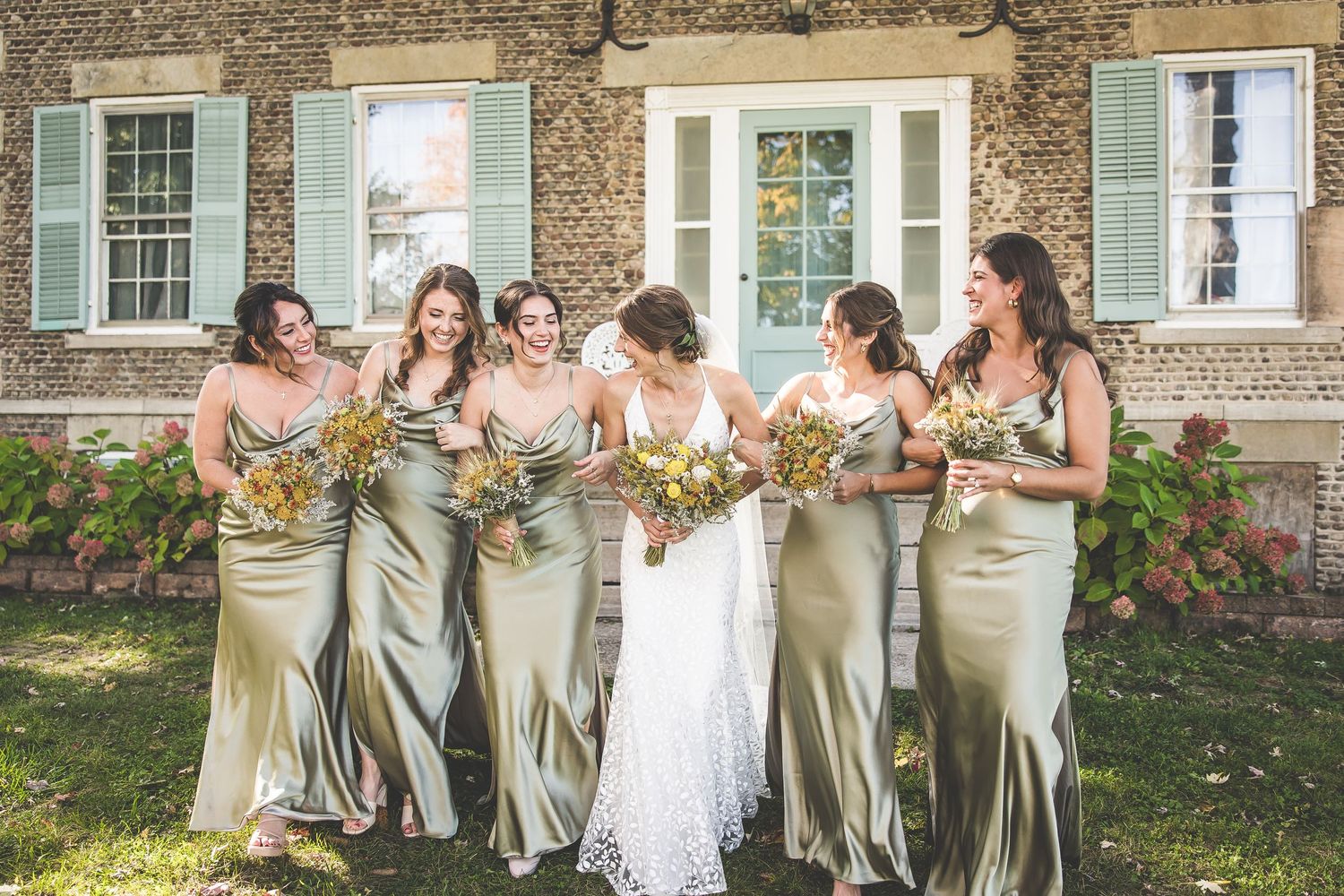 Wedding party in sage green dresses gather outdoors for group photo in front of historic stone building.