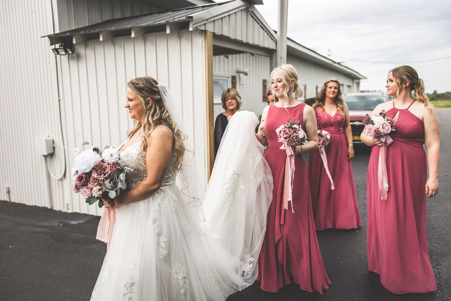 Bride in white gown stands with bridesmaids in rose dresses outside venue.