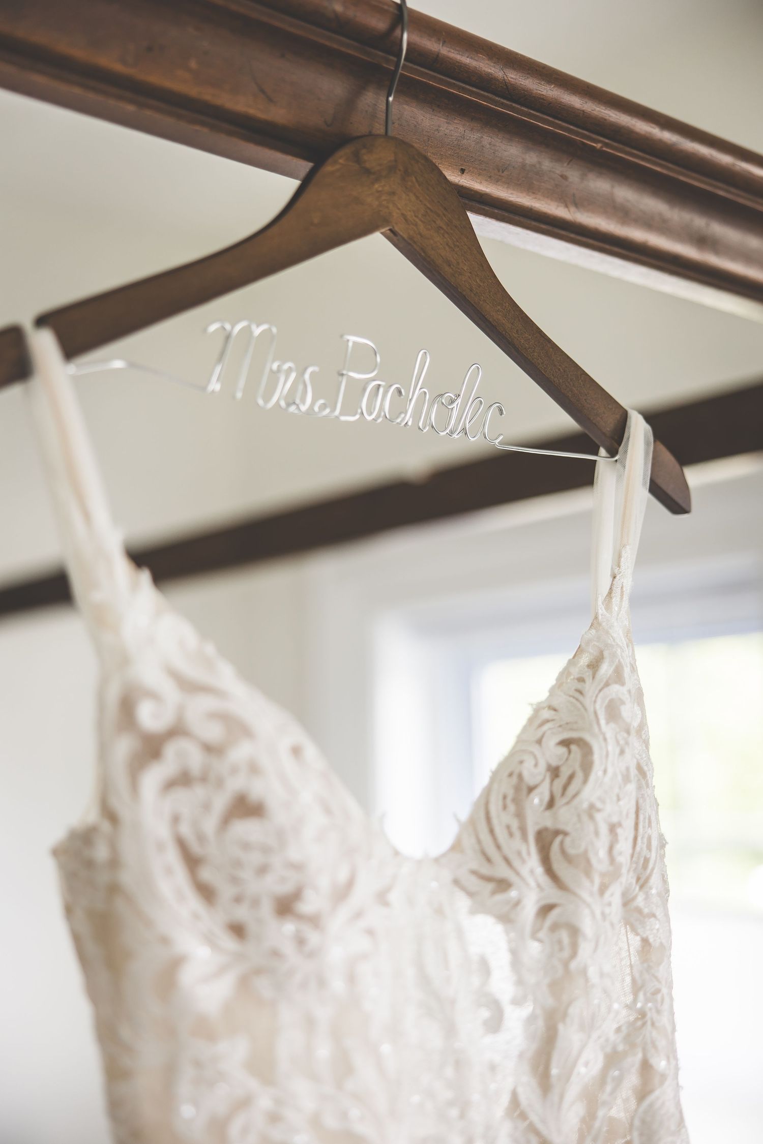 White lace wedding dress hanging on wooden hanger against light wall.
