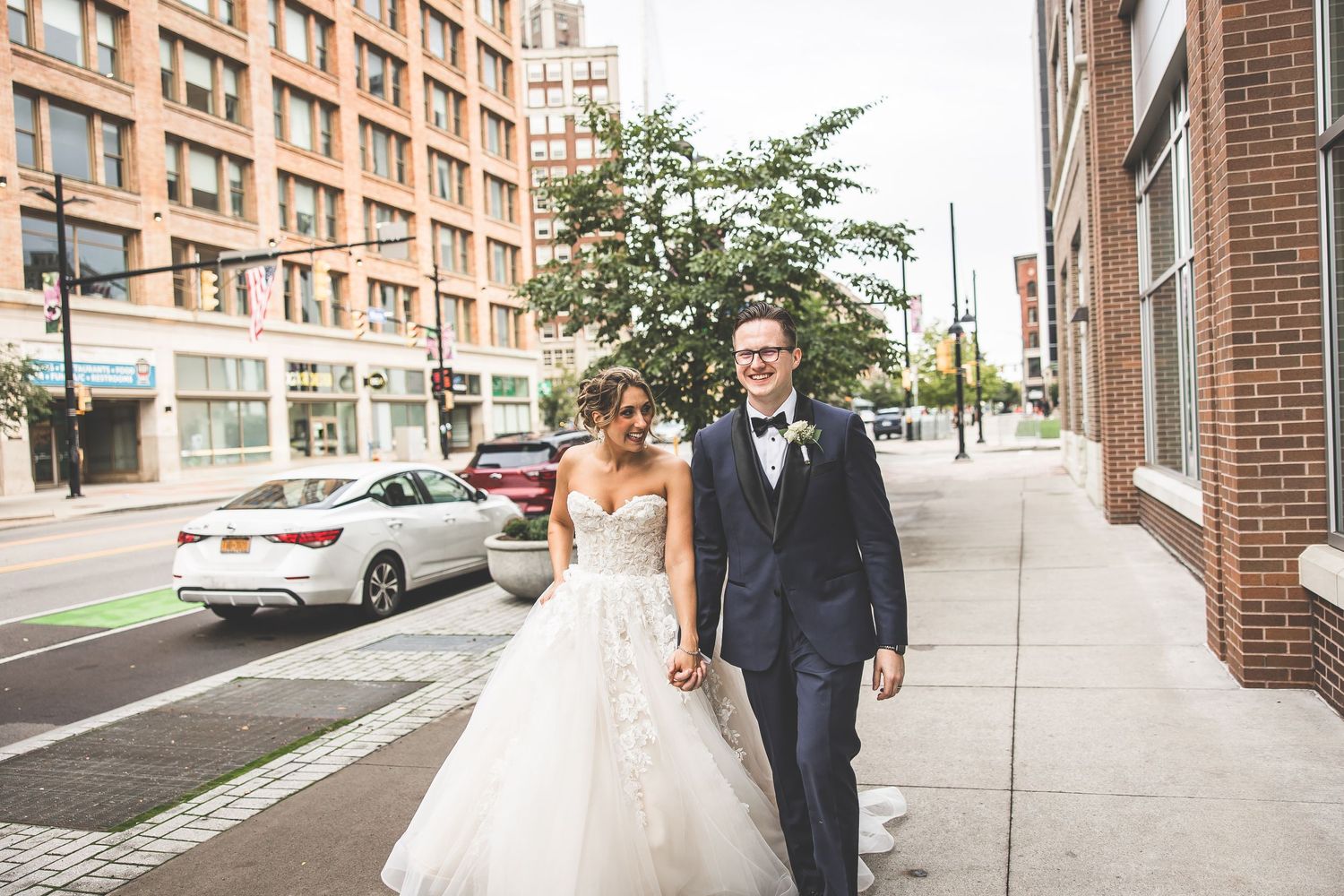 Newlyweds walking along a city street with modern architecture in the background.