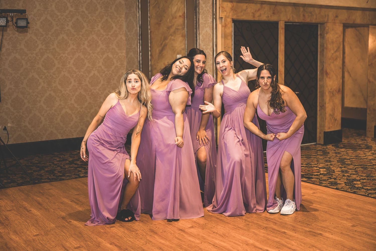 Bridesmaids in matching purple dresses pose playfully on a wooden dance floor.