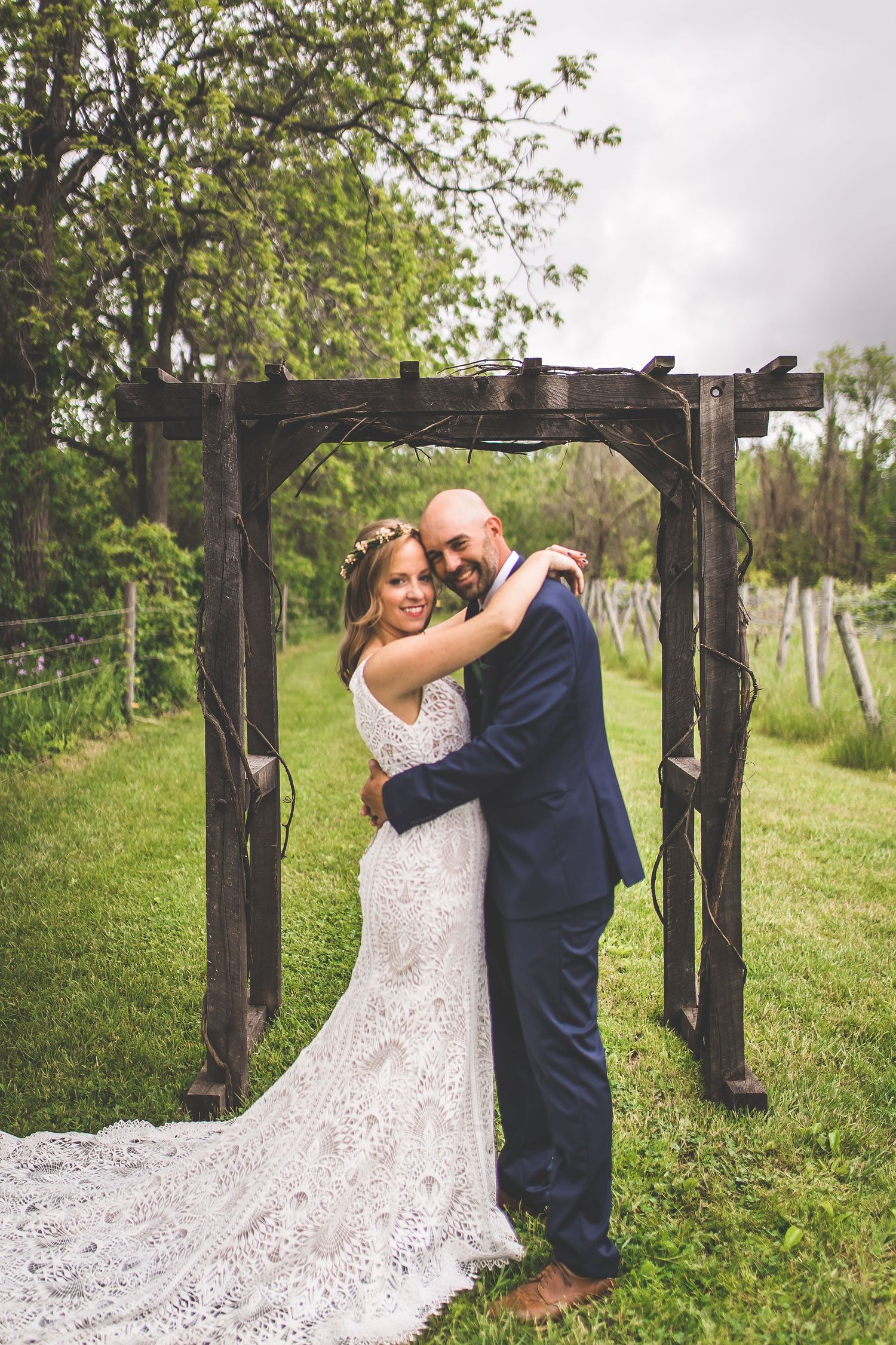 A romantic wedding photo under a rustic wooden arbor in a lush green garden setting.