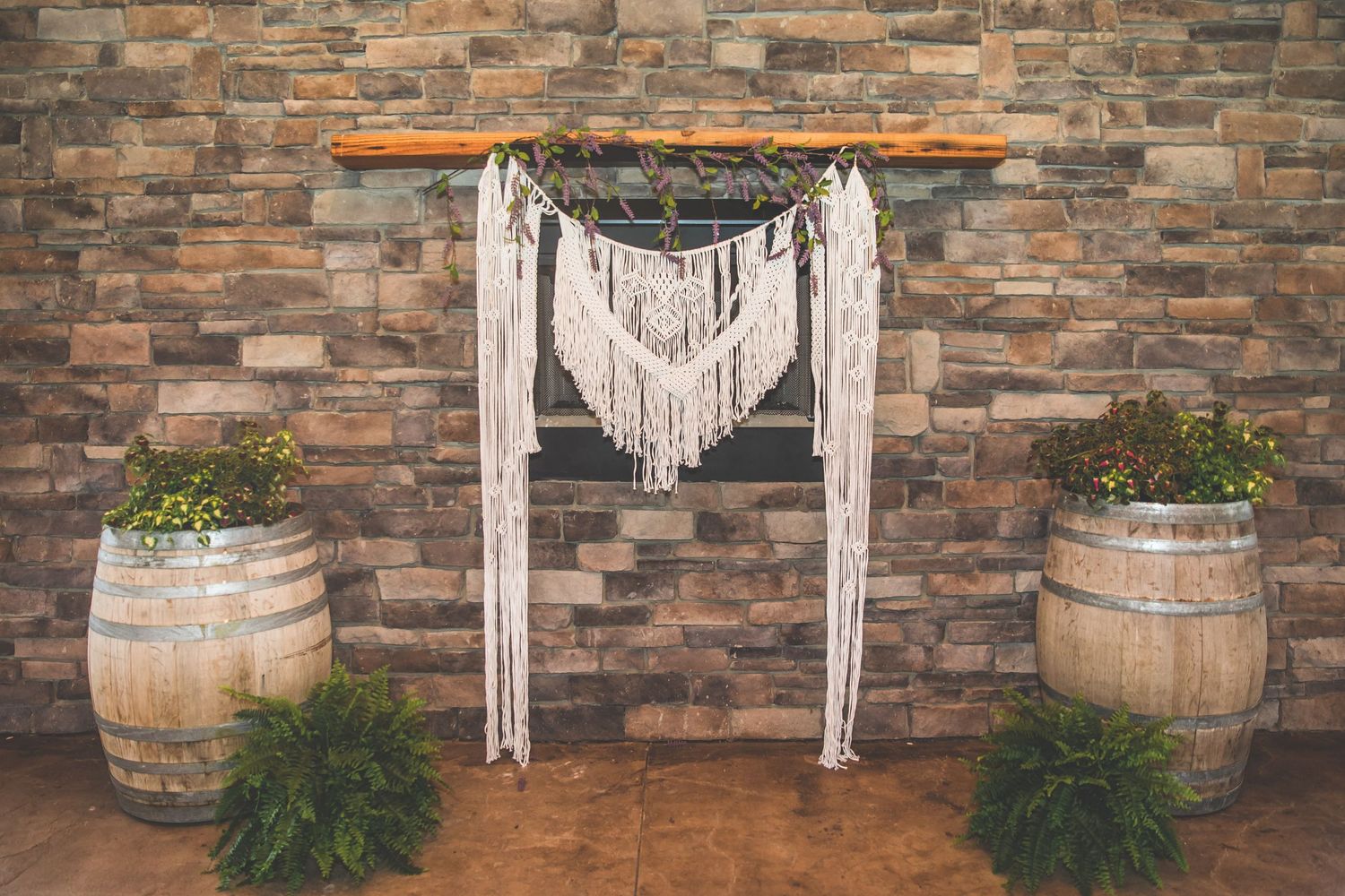 Stone wall backdrop with macrame hangings and wine barrels for wedding ceremony.