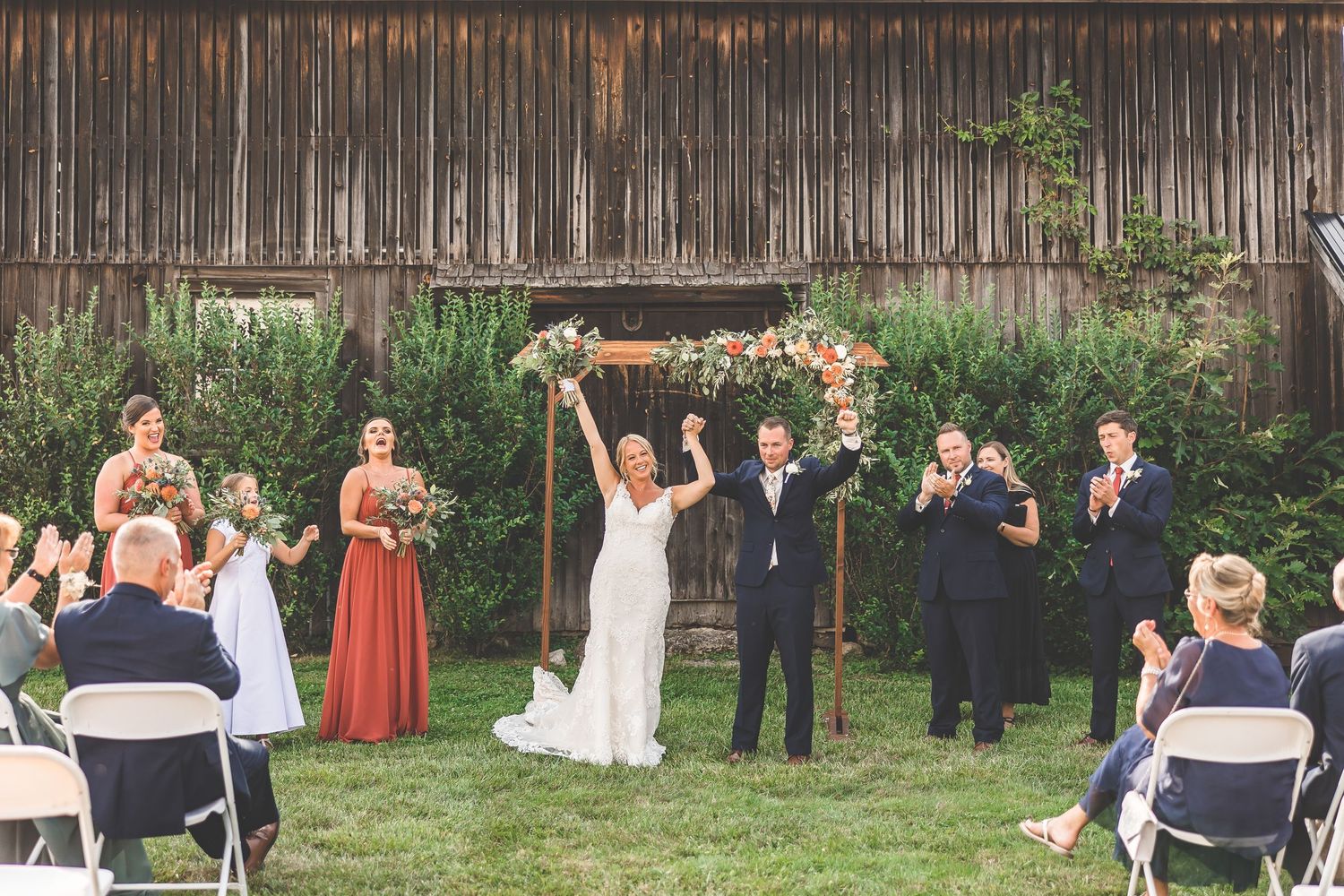 Wedding guests seated in white chairs on lawn watching outdoor ceremony at rustic venue.