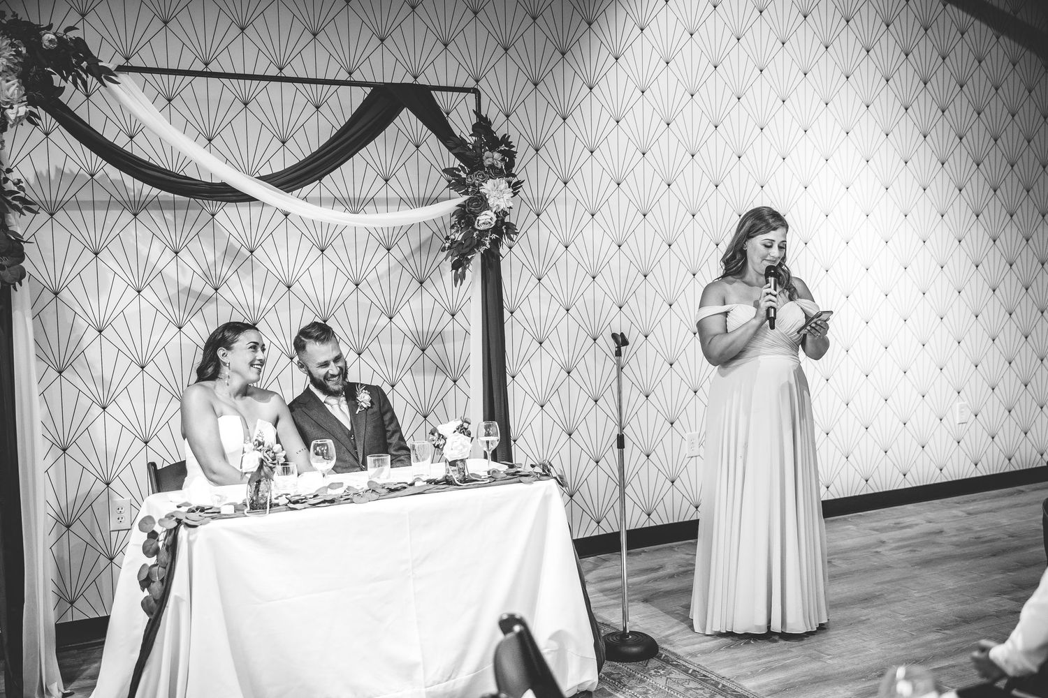 Black and white wedding reception photo with guest giving speech at formal event.