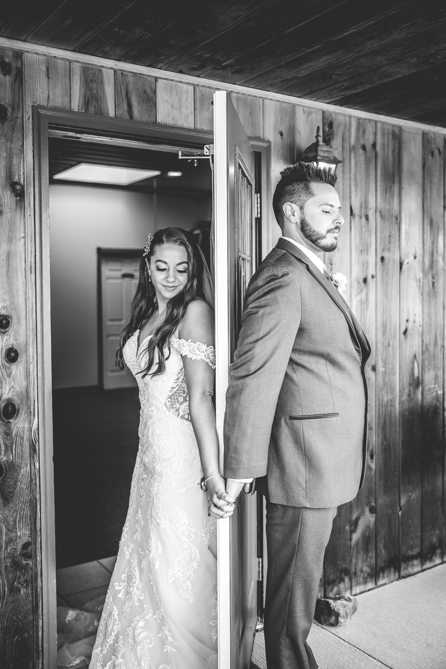 Couple shares a private first look moment while holding hands around a wooden door corner.