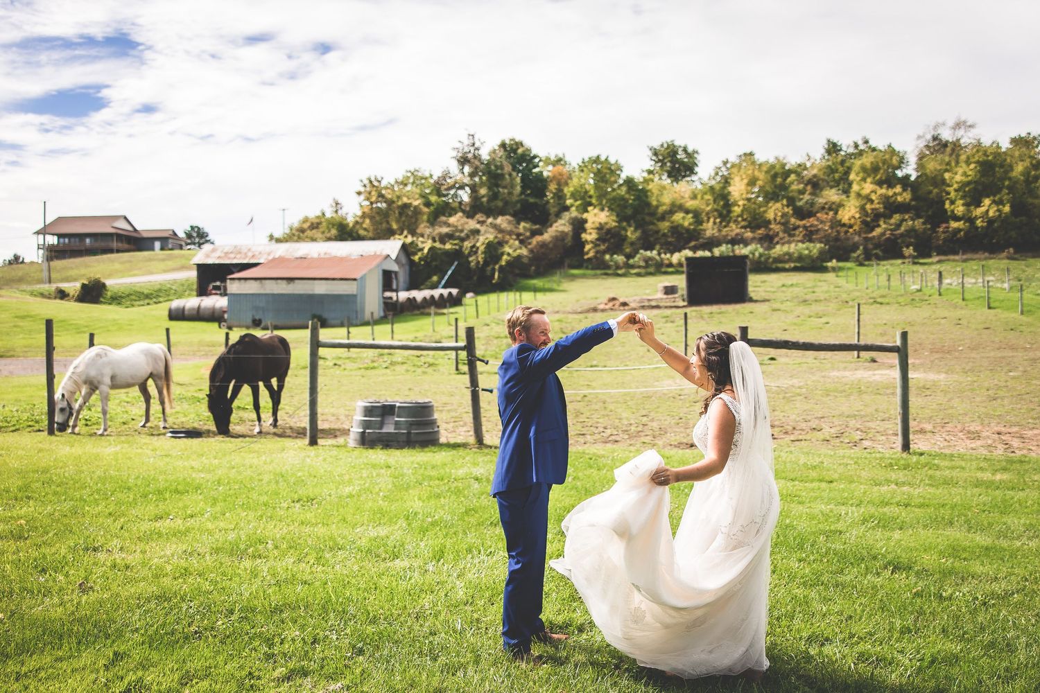 Newlywed couple walking and posing in a sunny outdoor field with rustic fencing and trees in the background.