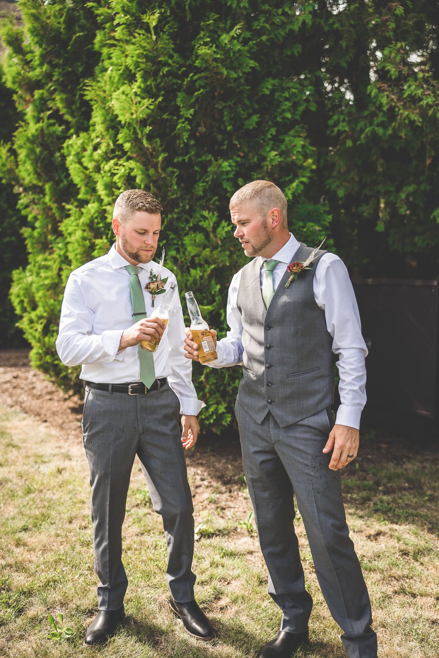 Groomsmen in gray vests and white shirts share drinks and laughs in an outdoor garden setting.