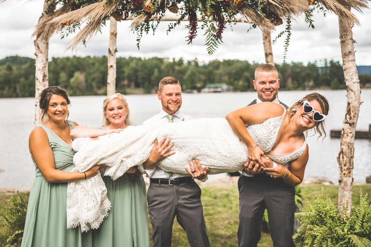 Wedding party members share joyful moments outdoors near birch trees and a lake.