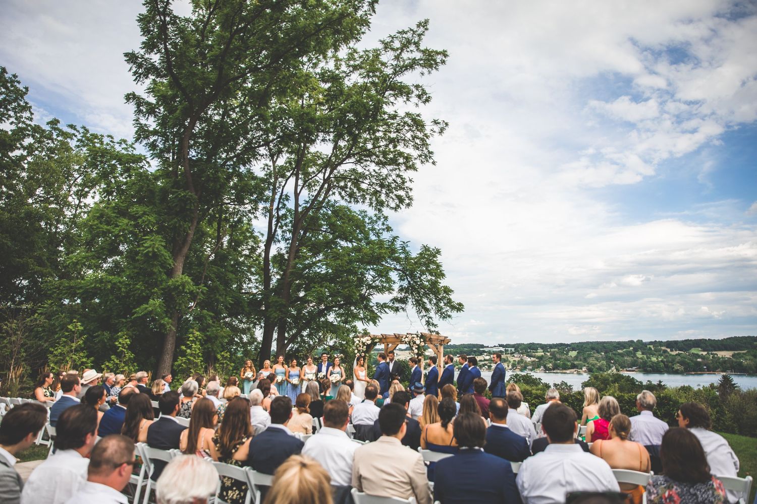 Panoramic view of an outdoor wedding ceremony overlooking a lake with guests seated on white chairs.