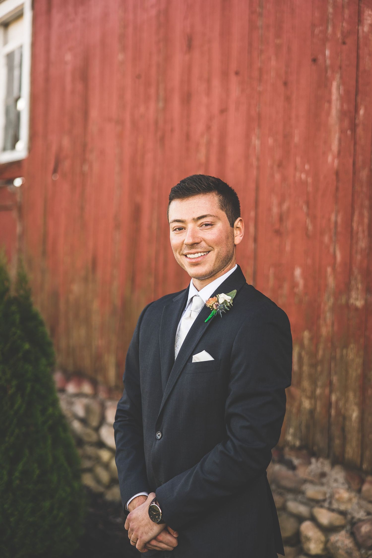 Groom in classic black tuxedo smiling against red barn background.