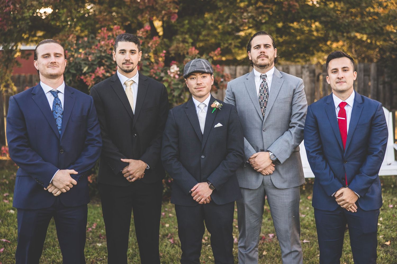 Groomsmen in formal suits stand in a row outdoors during an autumn wedding ceremony.