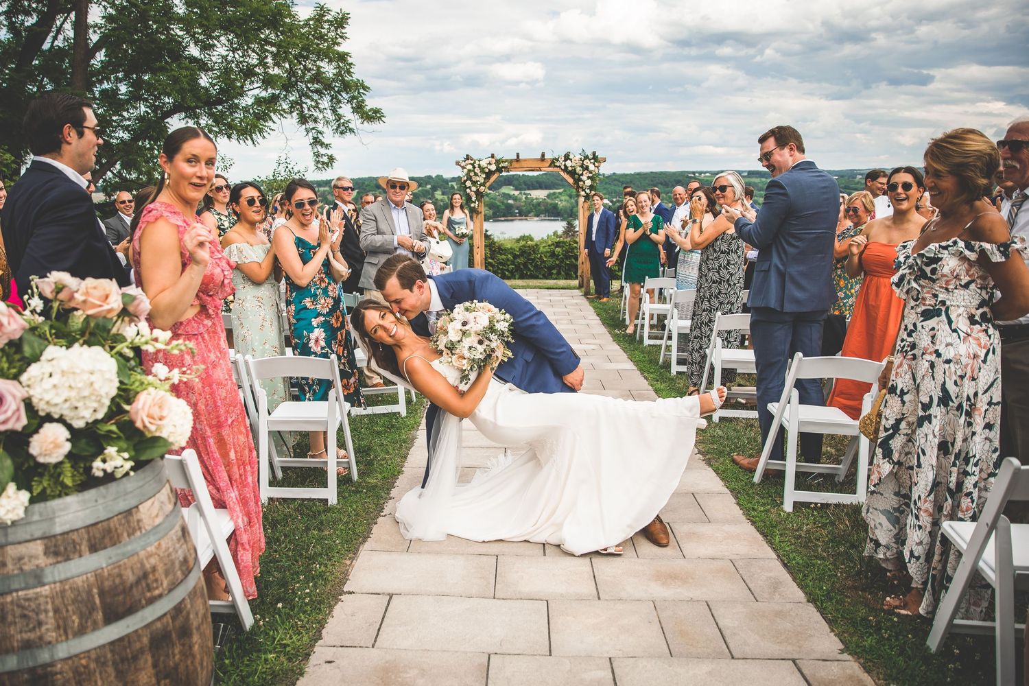 Wedding guests gather outdoors for a celebration on a beautiful summer day with lush greenery in the background.