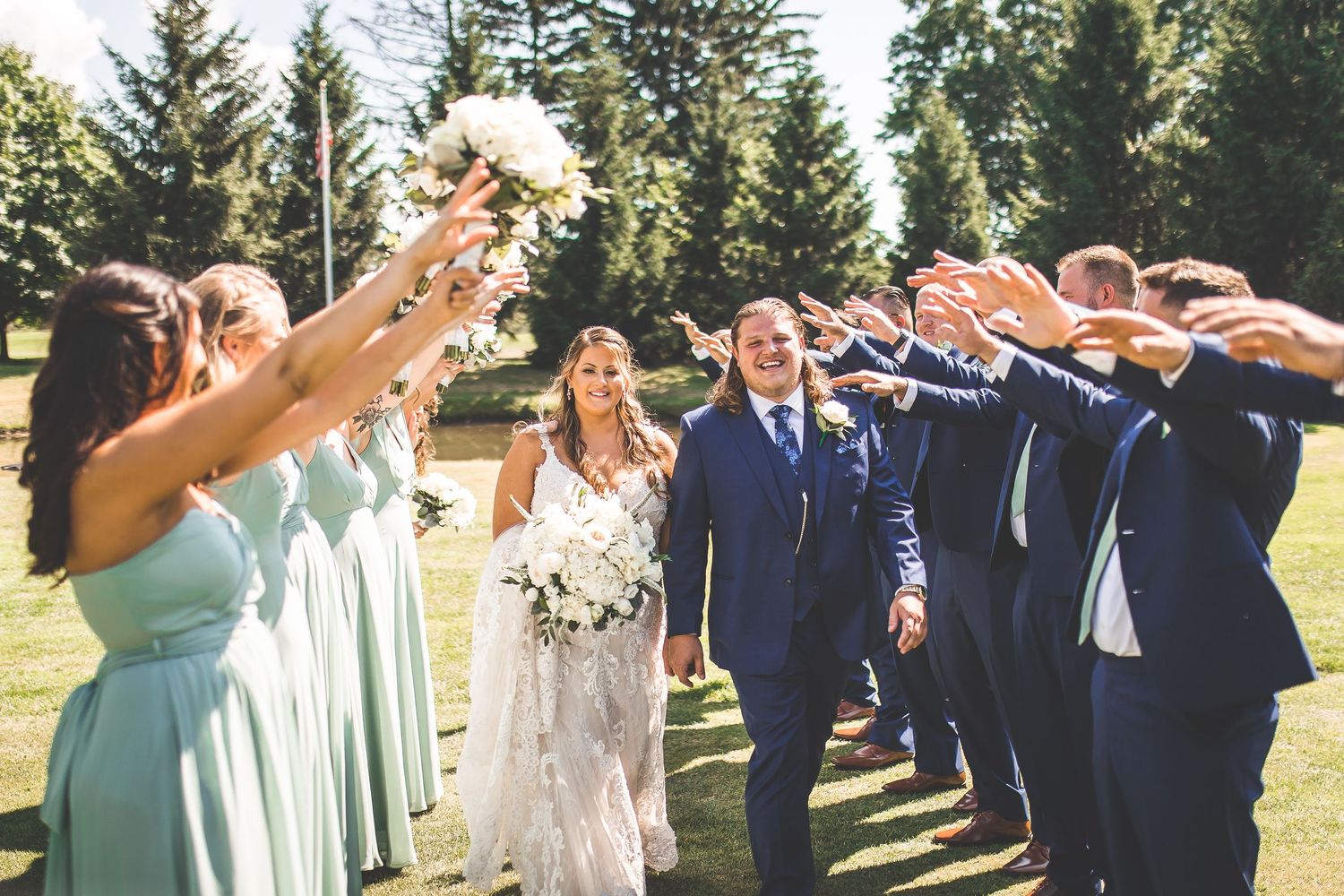 Wedding party poses joyfully on a sunny day with evergreen trees in the background.
