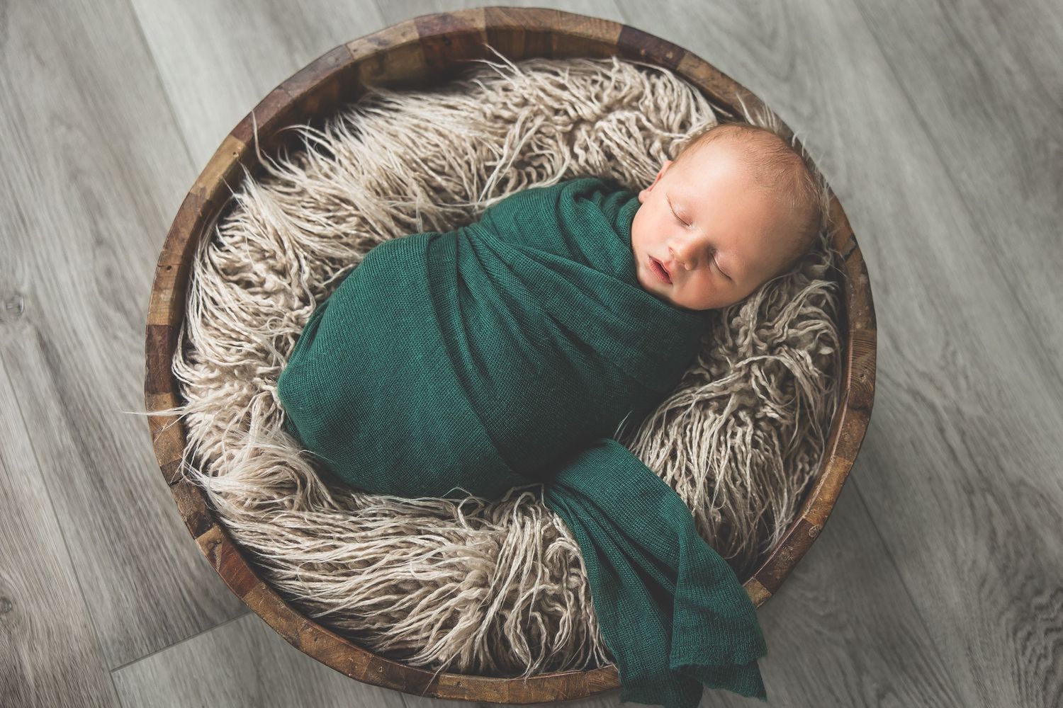 Sleeping newborn wrapped in teal swaddle resting in wooden bowl with fluffy gray fur lining.