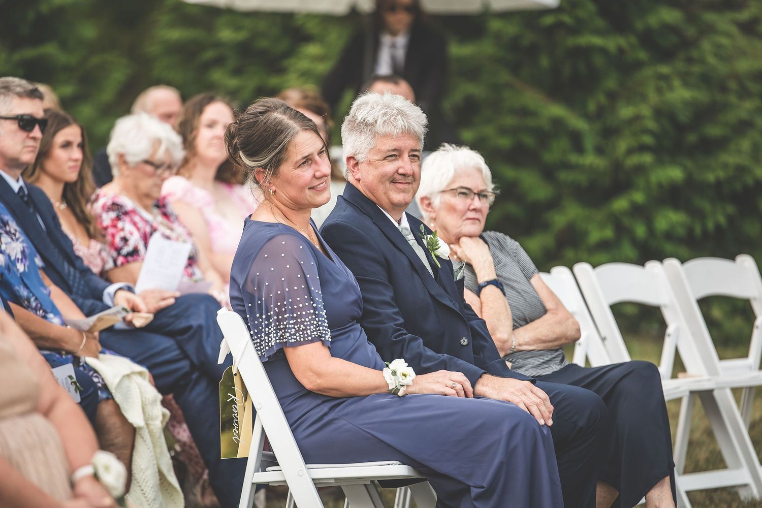 Wedding guests seated in white chairs watching outdoor ceremony.