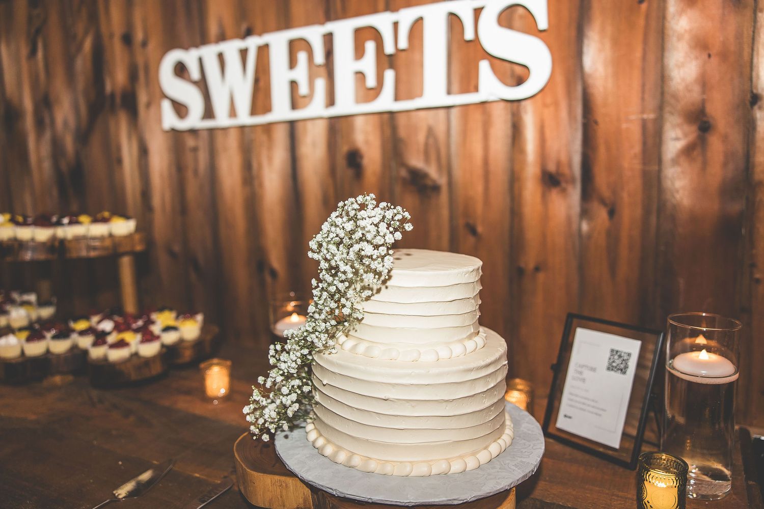 Rustic wooden wedding reception setup with white cake and 'SWEETS' signage.
