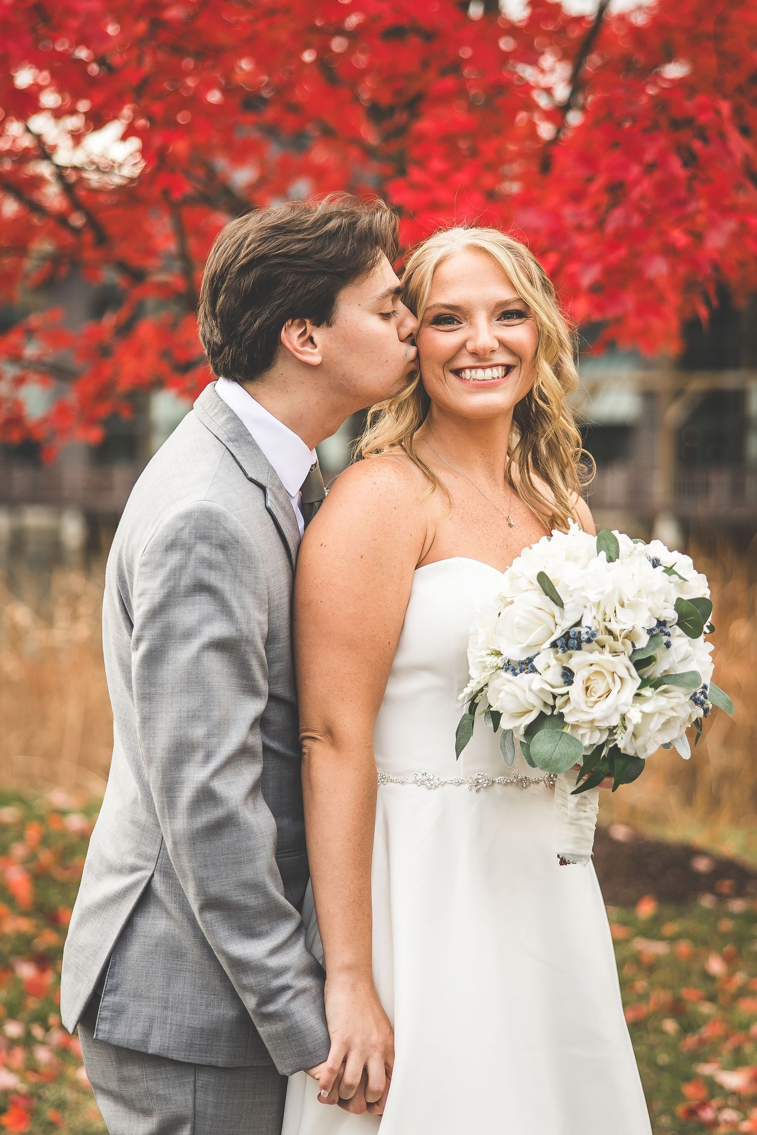 A romantic autumn wedding photo with vibrant red maple trees in the background and a white bouquet.