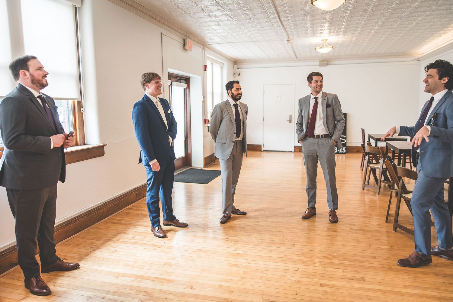 Wedding party members in formal attire gathering in bright hallway with hardwood floors.