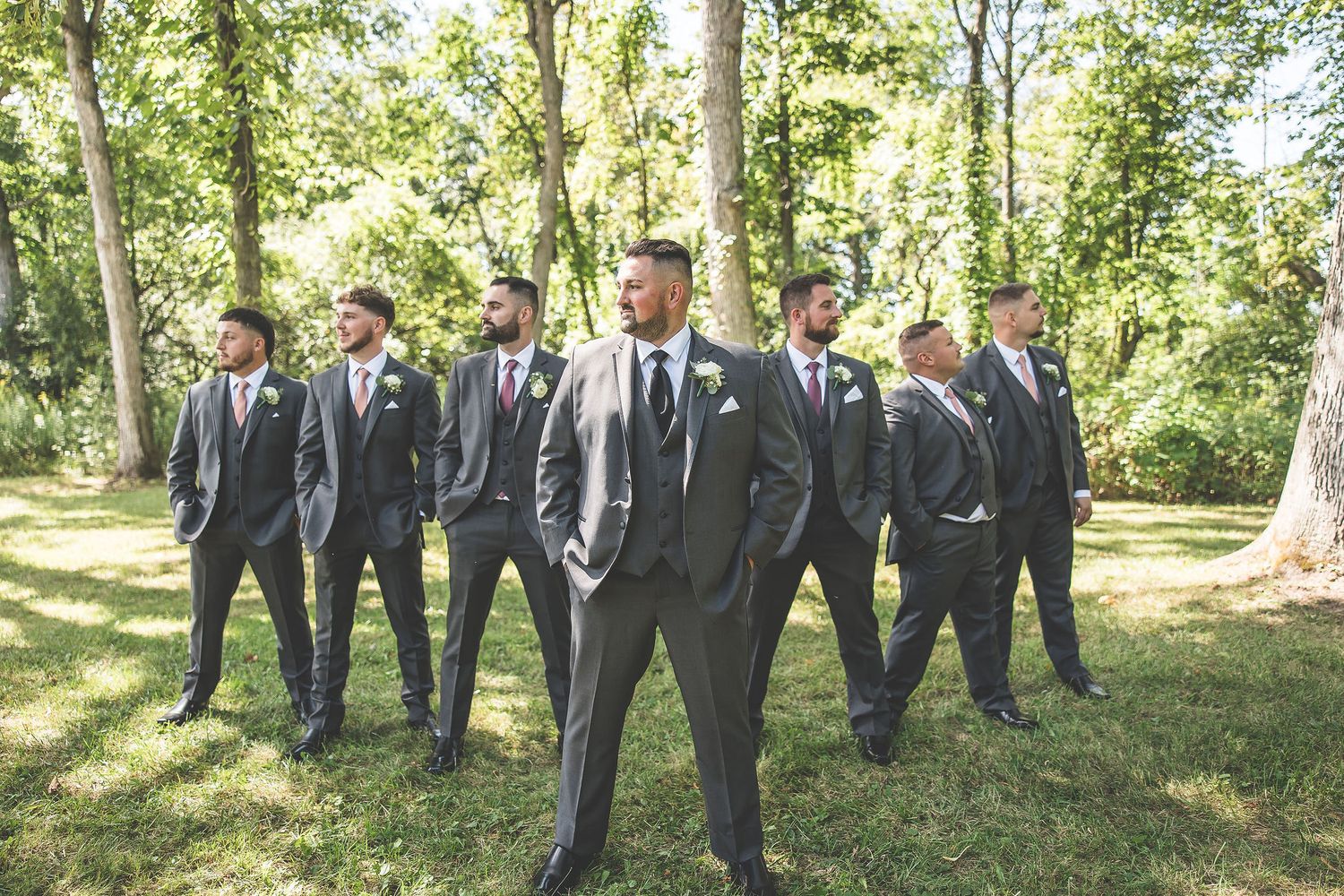 Groomsmen in formal attire pose casually in a line through a sunny wooded area.