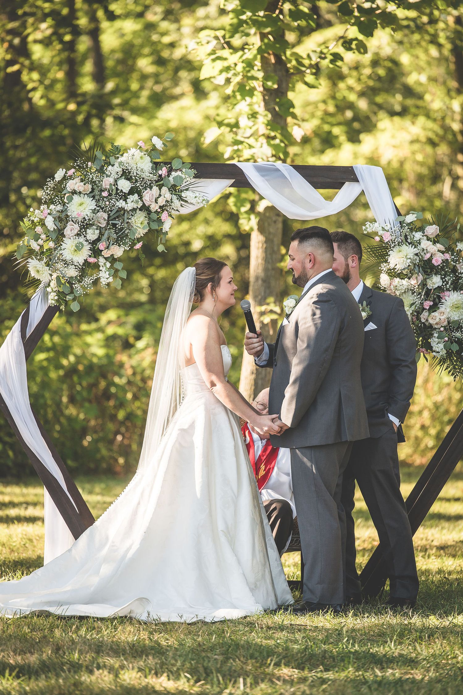 A wedding ceremony under a floral arch with white draping in a sunlit forest setting.