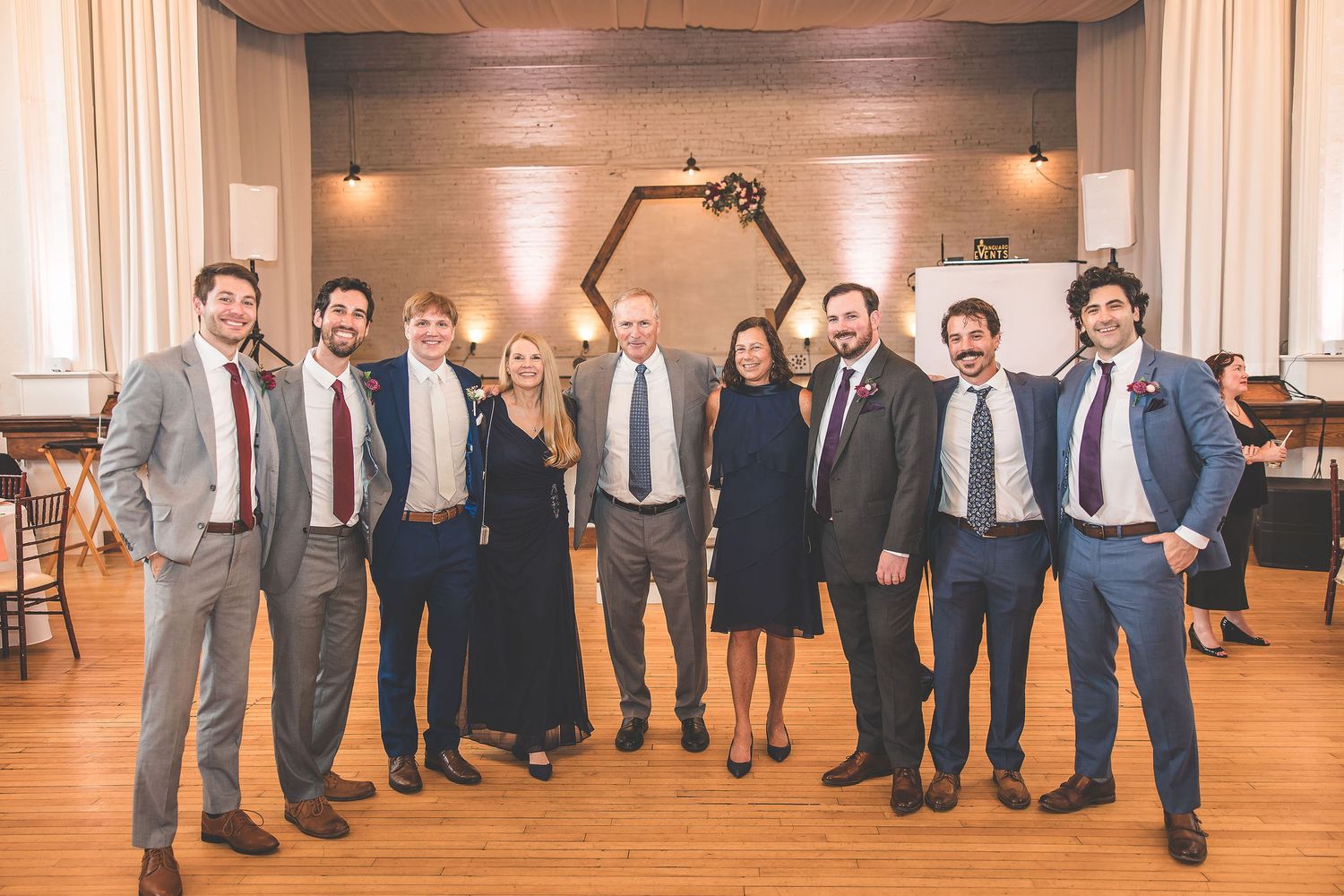 Wedding party members pose in formal attire against a light-colored backdrop.