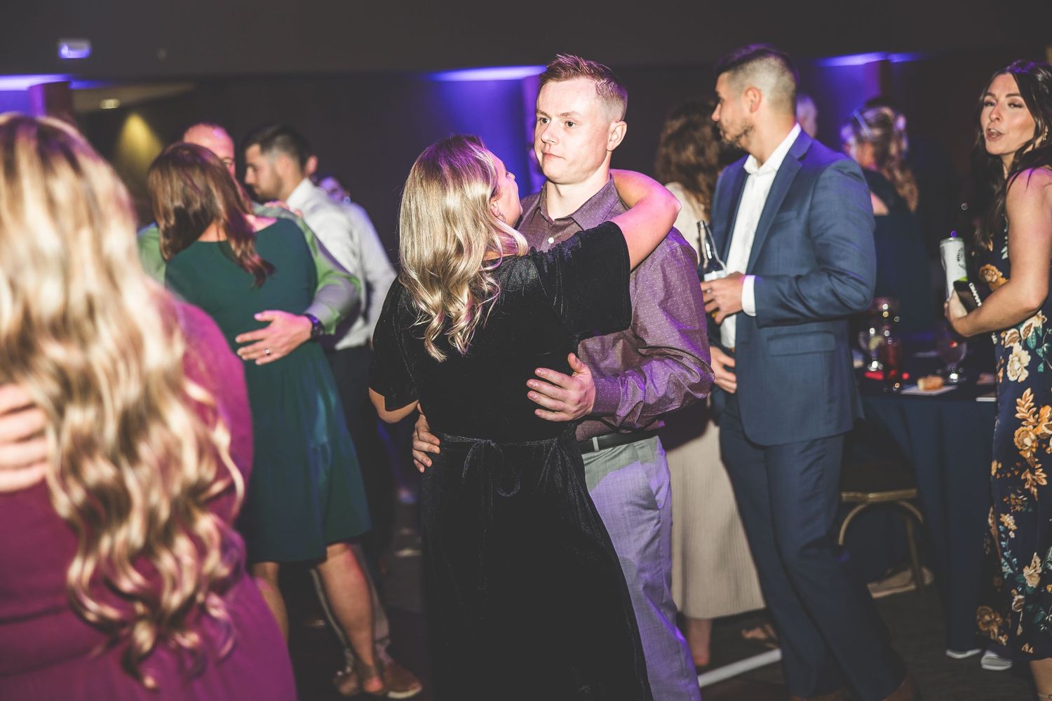 Crowded wedding reception dance floor with guests dancing under purple and blue lighting.