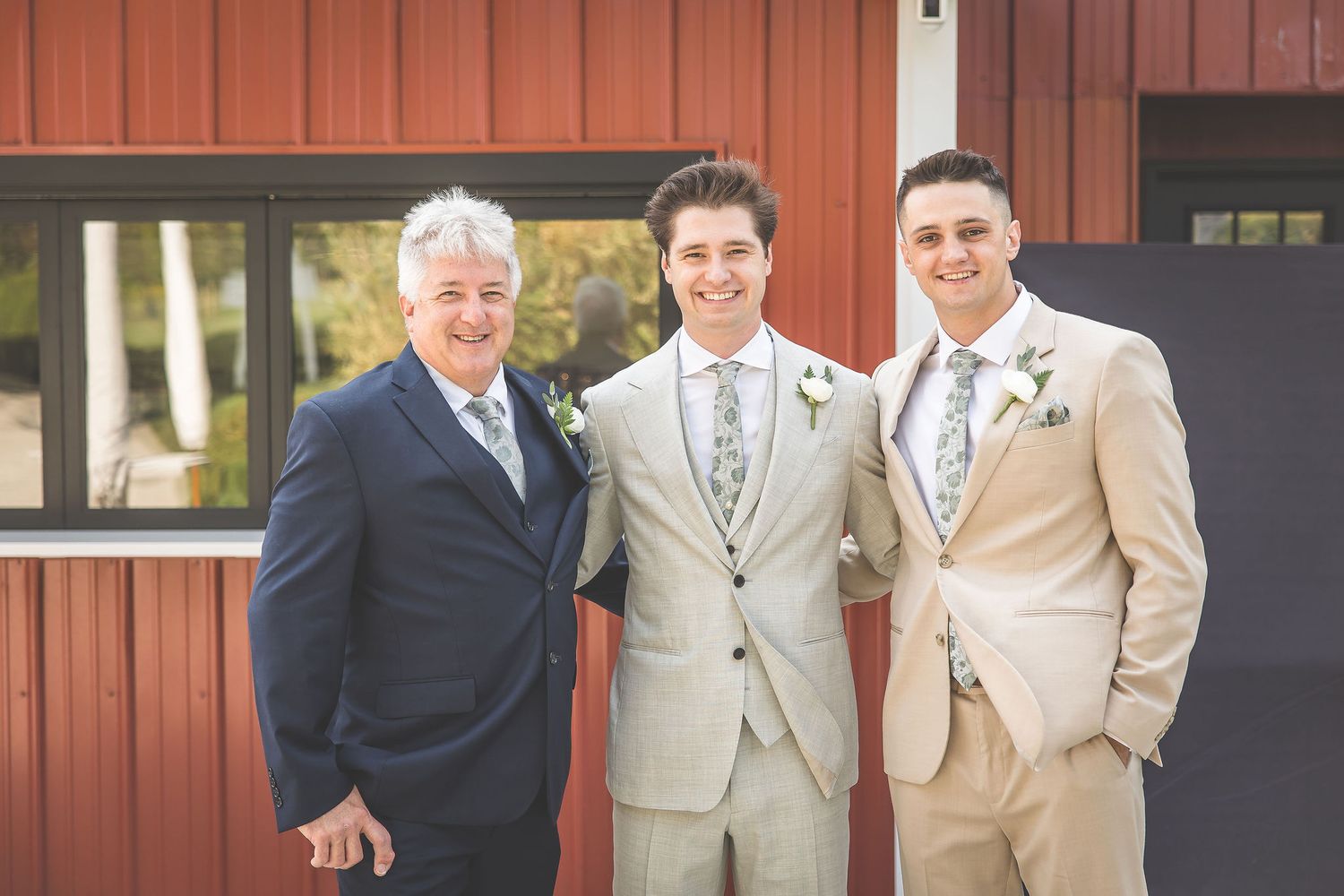 Wedding party members in tan suits and navy attire gather for photos by a rustic red barn.