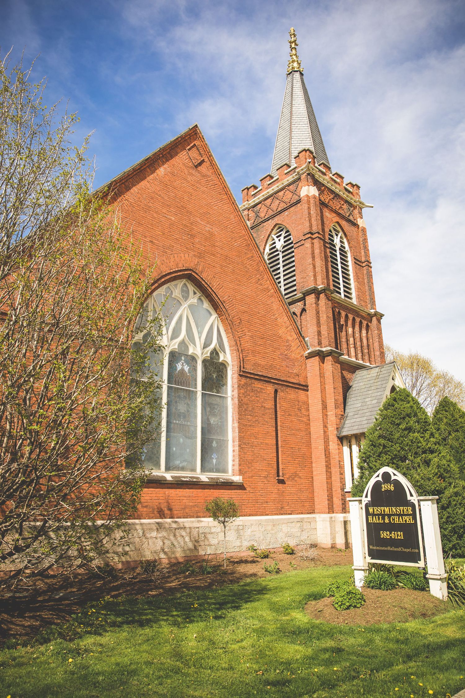Red brick church with Gothic windows and tall spire against blue sky.