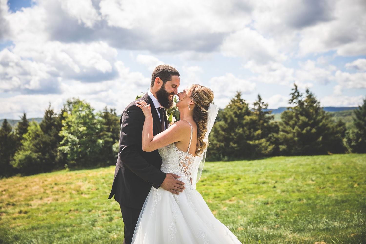 A romantic wedding couple embraces in a scenic mountain meadow with dramatic clouds overhead.