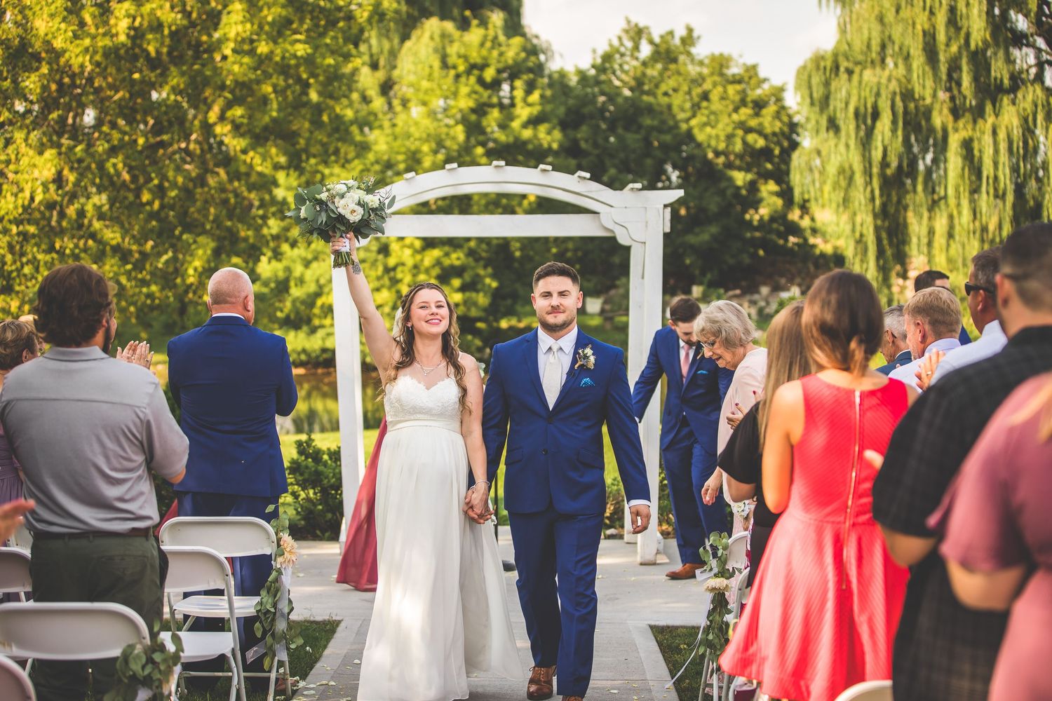 Wedding party poses under a white pergola surrounded by lush summer gardens and greenery.